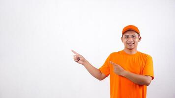 Smiling Asian deliveryman in orange t-shirt uniform poses as if he is pointing something in empty space area isolated on white background photo