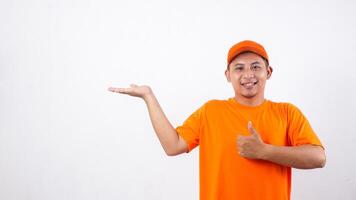 Asian deliveryman in orange t-shirt uniform poses as if he is presenting something in empty space area and showing thumb up isolated on white background photo