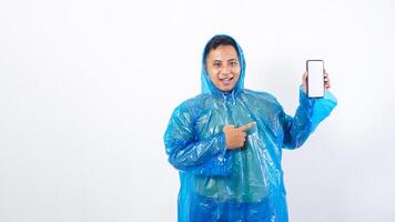 Portrait of Asian young man in blue raincoat pointing at smartphone mock up with blank screen isolated on white background photo