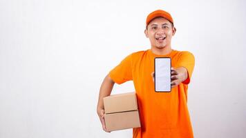 Smiling Asian Delivery man in orange t-shirt carrying package boxes parcel while showing a smartphone with blank screen isolated on white background photo