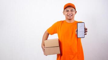 Portrait of Asian Delivery man in orange t-shirt carrying package boxes parcel while showing a smartphone with blank screen isolated on white background photo