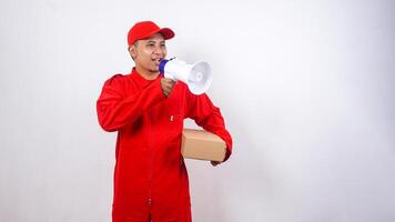 Portrait of Asian Delivery man in red uniform carrying a package box parcel while talk using megaphone isolated on white background photo