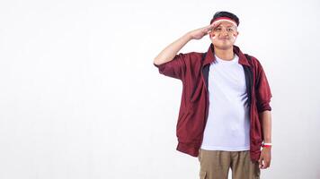Smiling Indonesian man with red and white ribbon wristband doing salute pose to celebrate independence day of Indonesia isolated on white background with copy space photo