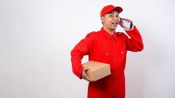 Portrait of Asian Delivery man in red uniform carrying a package box parcel while taking a phone call isolated on white background photo