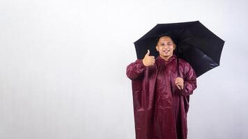 Smiling Asian man in red raincoat holds umbrella and showing his thumb up isolated on white background photo