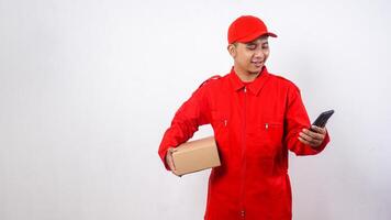 Asian Delivery man in red uniform carrying a package box parcel while looking at his smartphone screen isolated on white background photo