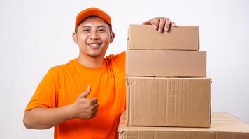 Smiling Asian deliveryman in orange t-shirt uniform with stack of package boxes parcel and showing his thumb up isolated on white background photo