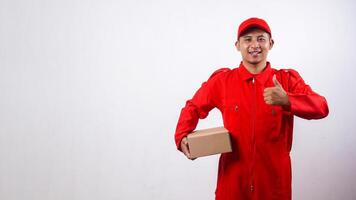 Delivery man in red uniform holding cardboard parcel box and showing thumb up isolated on white background photo
