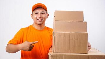 Smiling Asian deliveryman in orange t-shirt uniform with stack of package boxes parcel and pointing at the boxes isolated on white background photo