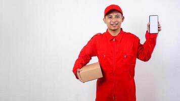 Portrait of Asian Delivery man in red uniform carrying a package box parcel while showing smartphone with blank screen isolated on white background photo