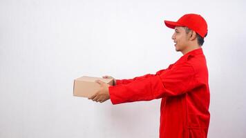 Side view of Asian Delivery man in red uniform poses as if he is giving package box parcel to customer isolated on white background photo