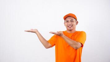 Smiling Asian deliveryman in orange t-shirt uniform poses as if he is presenting something in empty space area isolated on white background photo