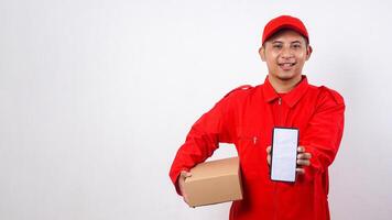Asian Delivery man in red uniform carrying a package box parcel while showing smartphone with blank screen isolated on white background photo