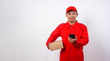 Asian Delivery man in red uniform carrying a package box parcel while holding a smartphone isolated on white background photo