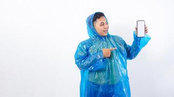 Portrait of smiling Asian young man in blue raincoat pointing at smartphone mock up with blank screen isolated on white background photo