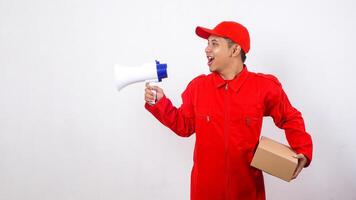 Portrait of Asian Delivery man in red uniform carrying a package box parcel while shout using megaphone isolated on white background photo