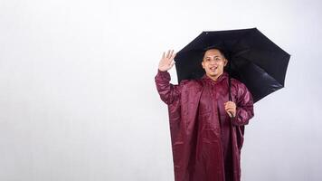 Smiling Asian man in red raincoat holds umbrella raising his hand to give greeting pose isolated on white background photo