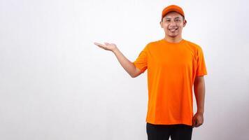 Smiling Asian deliveryman in orange t-shirt uniform poses as if he is presenting something in copy space area isolated on white background photo