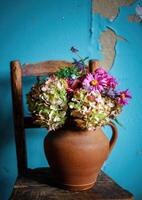 Bright and colorful flower arrangement in a clay pot on a rustic wooden chair against teal wall photo