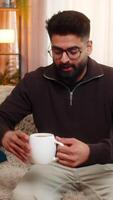 Indian man enters room on sofa enjoys coffee with a smile after stressful day at home with comfort video