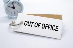 Out Of Office text on white notepad with alarm clock on white background. Away from office concept photo