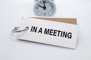 In A Meeting text on white notepad with alarm clock on white background. Away from office concept. photo