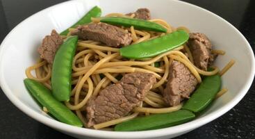 Delicious beef chow mein with crisp snow peas served in a ceramic bowl on dark counter photo