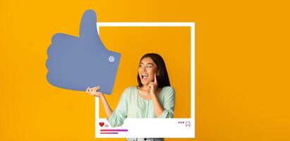A young woman joyfully poses with a large thumbs-up symbol against a vibrant orange background, expressing enthusiasm for her online content. This moment highlights her connection with followers. photo