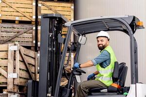 Employee on electric forklift carry container with ripe apples at storage. Smiling millennial bearded man drives in hard hat in truck, lifts up fruit, on many wooden crates at warehouse, free space photo