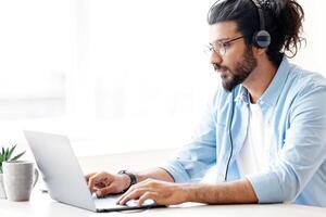 Handsome eastern freelancer guy working remotely with laptop at home office, using computer for online project or education, typing on keyboard, sitting at desk near window, copy space photo