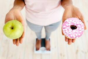 Healthy vs unhealthy food. Black woman holding apple and donut on scales, making choice of her meal, above view. African American lady deciding on her diet, choosing balanced nutriton photo