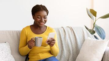 Domestic Relax. Positive black lady resting on couch with smartphone and coffee, browsing internet and enjoying hot drink, african woman checking social networks or messaging with friends, copy space photo