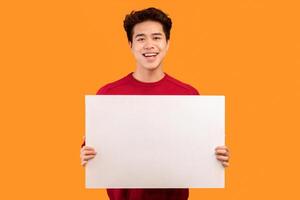 Advertisement Concept. Portrait of smiling asian man holding empty blank board isolated on orange studio background. Happy guy standing with white square paper placard for ad, showing it to camera photo
