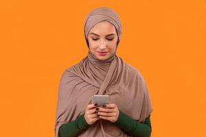 Muslim Lady Using Cellphone Mobile App Or Texting Networking In Social Media Online Standing Over Yellow Background. Phone User, New Application Advertisement Concept. Studio Shot photo