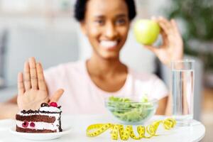 Happy black woman choosing fresh apple over unhealthy cake, refusing to eat dessert at home, selective focus. Young African American lady preferring fruit over junk food. Copy space photo