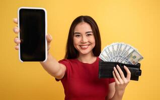 A cheerful young woman holds a smartphone in one hand and a cash-filled wallet in the other, smiling against a bright yellow background, representing financial success and modern tech. photo