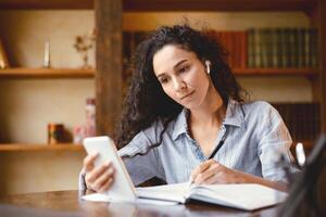 Remote Learning. Portrait of young woman in wireless earbuds sitting at desk, using smart phone and writing in her notebook, taking notes, watching lecture, doing homework, studying online at home photo