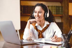 Doing Homework. Portrait of smiling young woman sitting at desk with pen, writing and taking notes in notebook, using and looking at laptop computer. Cheerful curly lady watching online course photo