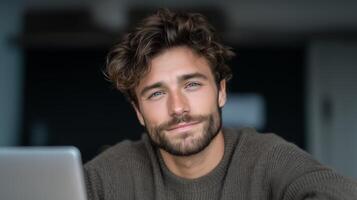 A man with curly hair and a beard smiles while using a laptop in a contemporary workspace. He interacts with a voice assistant, showcasing a seamless connection for communication and productivity. photo