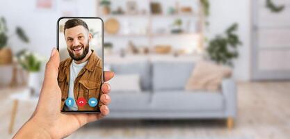 A person is holding a smartphone during a call. The background features a comfortable living room with a sofa, plants, and decorative shelves, creating a relaxed atmosphere. photo