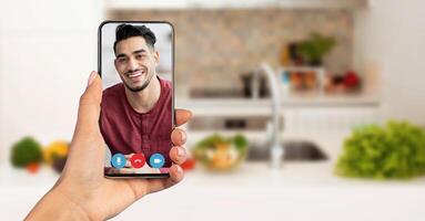A person is using a smartphone to connect with a friend on a call. The background features a modern kitchen filled with fresh vegetables, creating a warm home atmosphere. photo