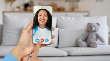A person is holding a smartphone with smiling woman on screen, have an online conversation. The setting is a cozy living room with a teddy bear on the couch, creating a warm atmosphere. photo
