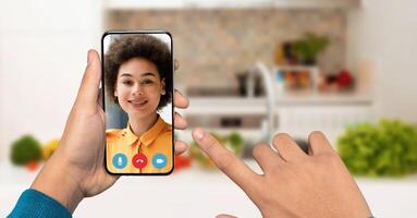 A hand holds a smartphone displaying a smiling person on the screen during a call. The background shows a bright kitchen with fresh vegetables, creating a lively atmosphere. photo
