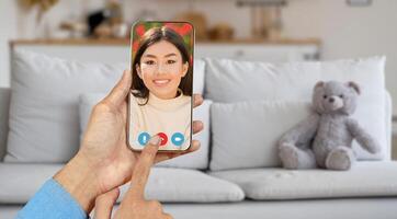 A person holds a smartphone showing a smiling individual on the screen, engaged in a call from a cozy living room with a teddy bear on the couch. photo