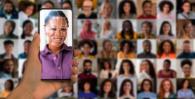 A person holds a smartphone in hand displaying a face scan. Behind, a diverse collage of people highlights the technology's ability to identify and recognize various faces in modern society. photo