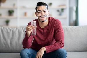 A man sits comfortably on a couch, smiling while holding a glass of water. His facial features are highlighted by recognition technology, showcasing modern advancements in identification. photo