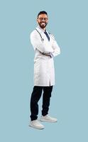 Middle-Eastern Male Doctor Posing Wearing White Uniform Crossing Hands Over Blue Background, Smiling To Camera. Full Length Shot Of Professional Medical Worker, Successful Physician photo