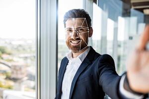 A man smiles as he uses facial recognition technology for identification in a contemporary office. Bright natural light floods the space, showcasing modern architecture and technology. photo