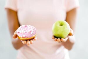 Choice of healthy or junk food. Closeup view of black woman choosing between donut and apple on light background. Millennial lady making decision about her snack, facing temptation photo