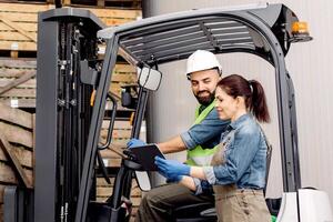 Management of loading products, modern device and checking driver work in warehouse. Cheerful male in helmet lifts up apples in container on shelves with wooden crates, lady control man on fork lift photo
